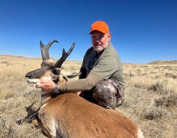 A hunter kneels beside a mature pronghorn antelope, gently holding its horns. The scene is set on an expansive prairie with a bright blue sky overhead.